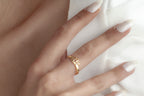 A close-up of a hand with white manicured nails wearing a delicate gold ring with a small floral or leaf motif, resting against a white textured fabric.