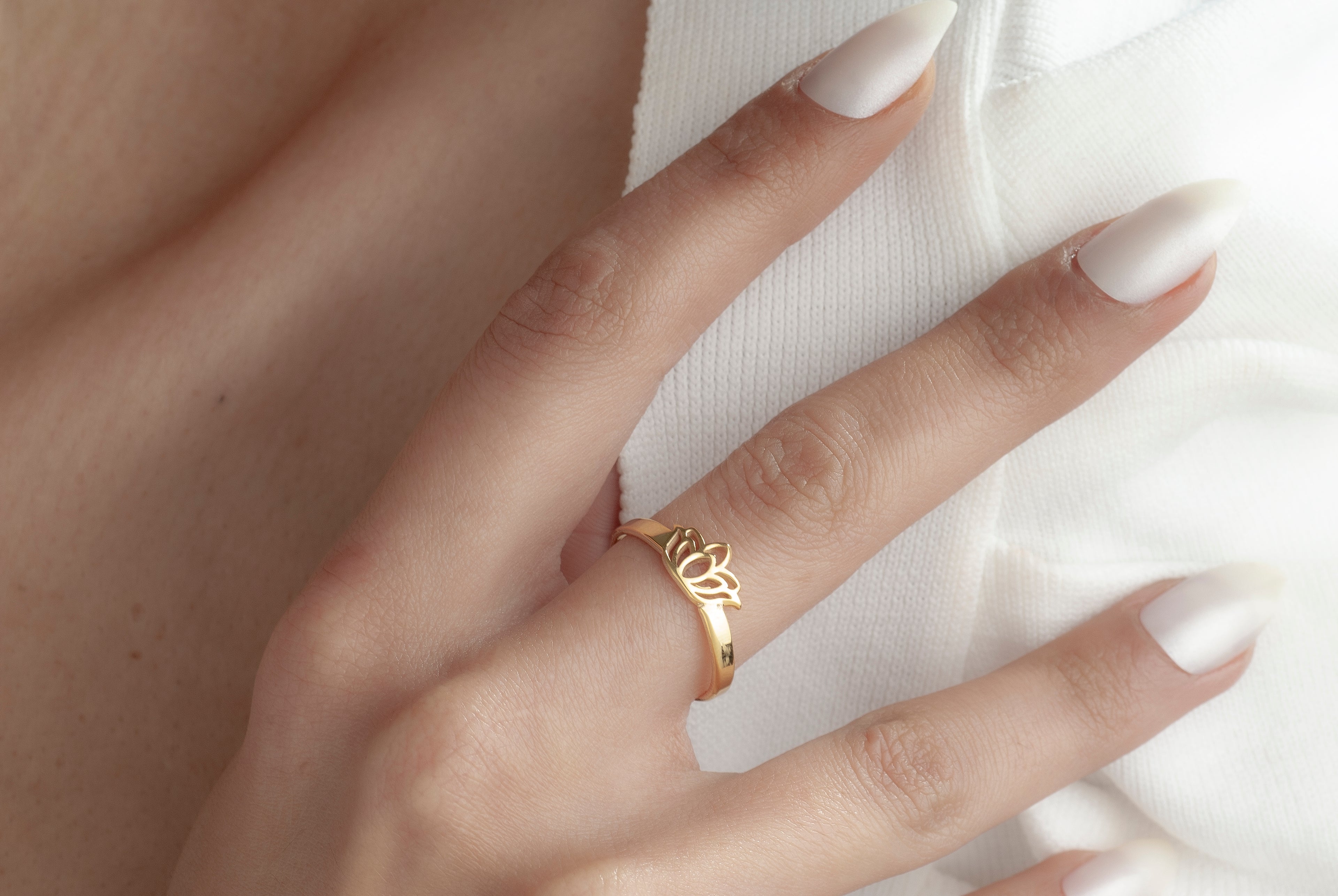 A close-up of a hand with white manicured nails wearing a delicate gold ring with a small floral or leaf motif, resting against a white textured fabric.